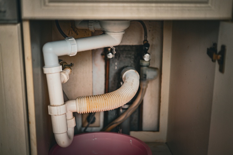Water dripping from under-sink pipe into bucket showing plumbing leak inside cabinet